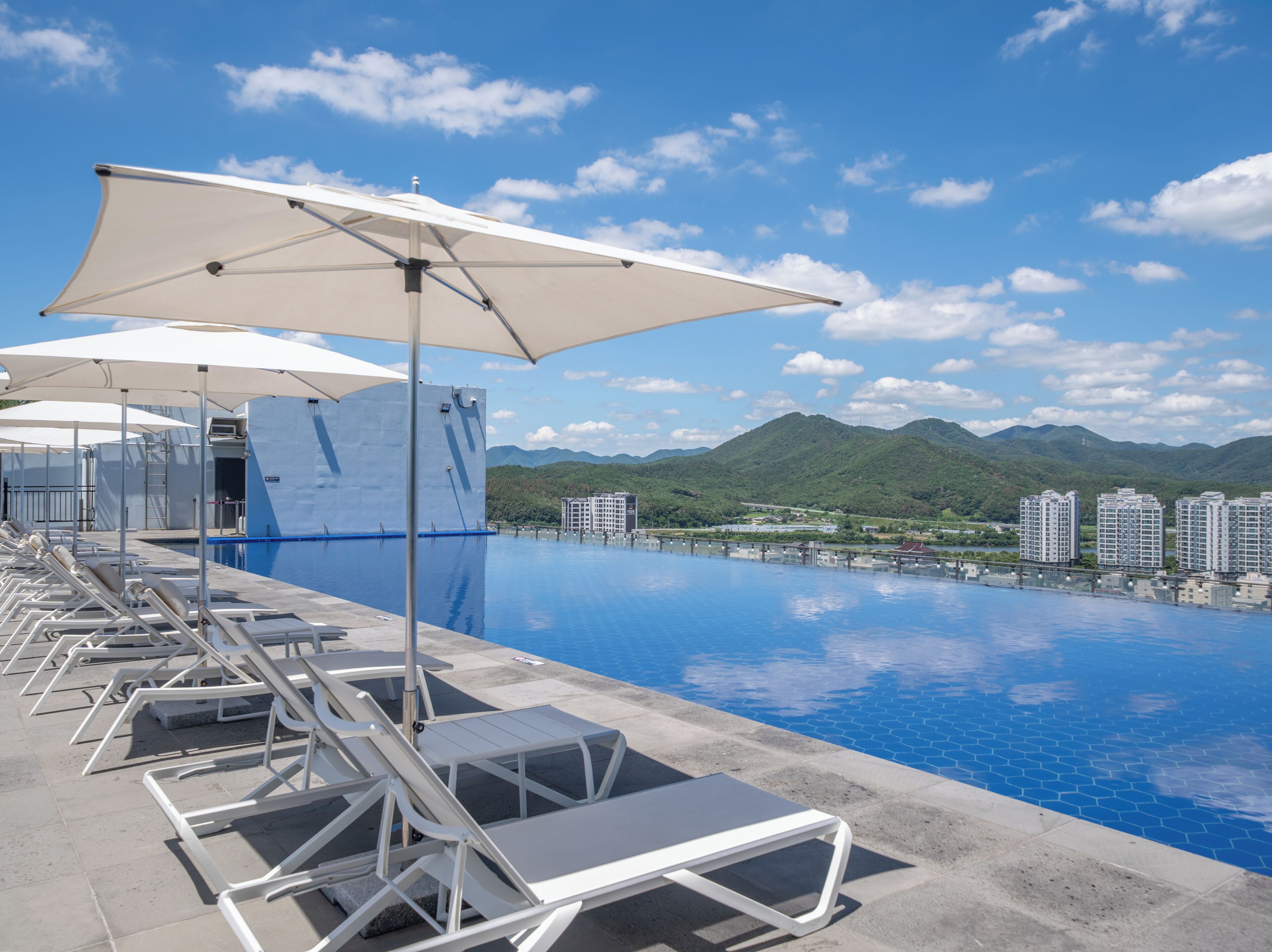A swimming pool with lounge chairs and umbrellas, surrounded by mountains and buildings.