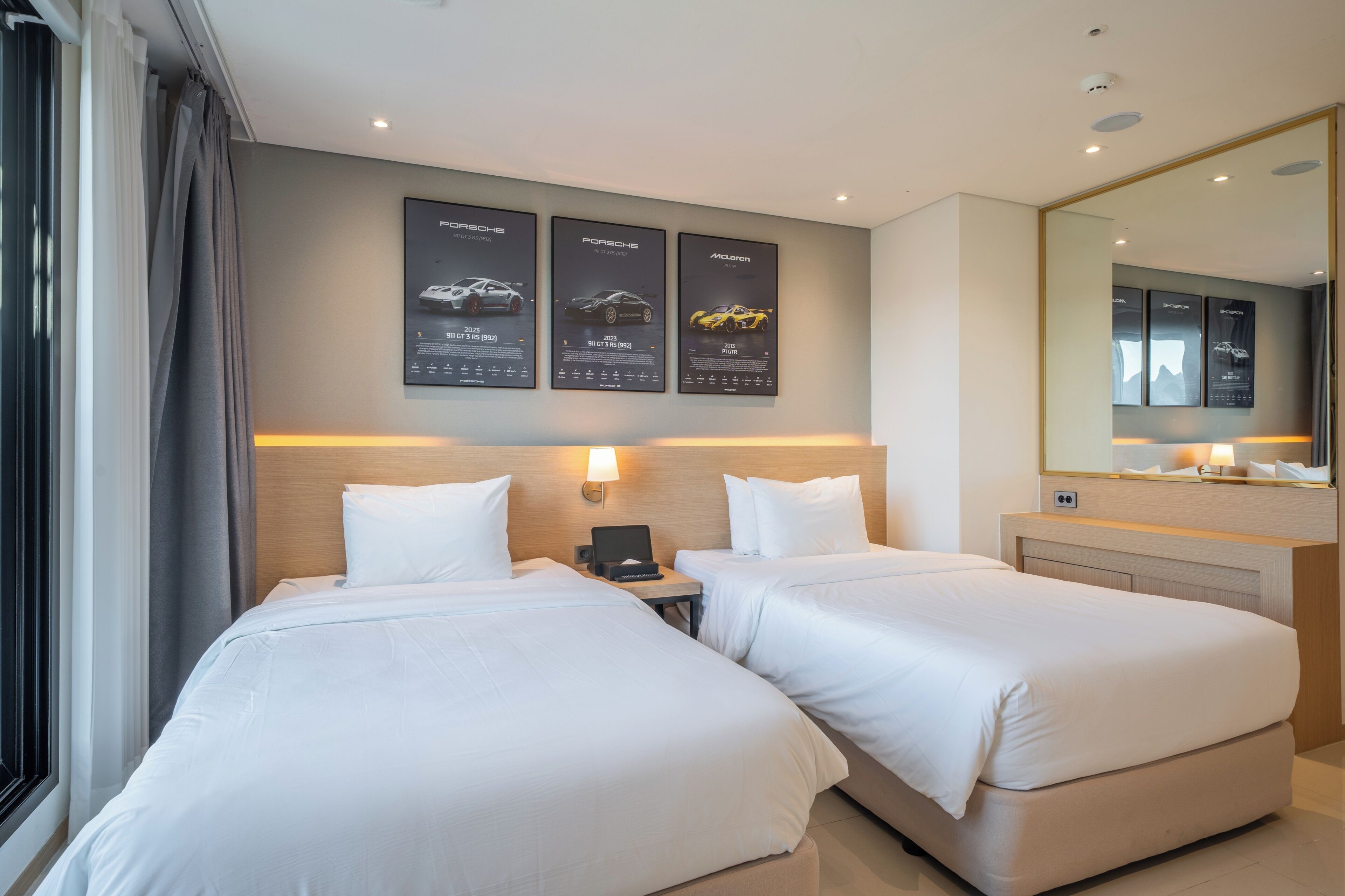 A hotel room with two beds, a nightstand, a lamp, a mirror, and wall-mounted posters of cars.