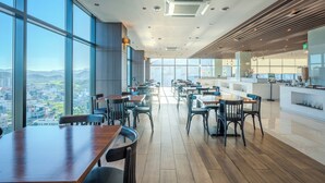 A modern restaurant with large windows offering city views, wooden tables and chairs, and a clean, contemporary interior design.