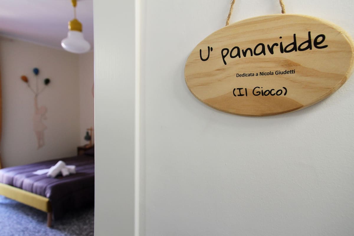 Un letrero de madera con el texto "U' panaridde" y "Dedicado a Nicola Giudetti (Il Gioco)" en una habitación con una cama y una decoración de pared.