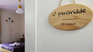 Uma placa de madeira com os textos "U' panaridde" e "Dedicato a Nicola Giudetti (Il Gioco)", em um quarto com cama e um ornamento na parede.