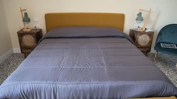Uma cama com cabeceira cor mostarda e roupa de cama cinzenta, ladeada por mesas de cabeceira de madeira com luminárias e alto-falantes.