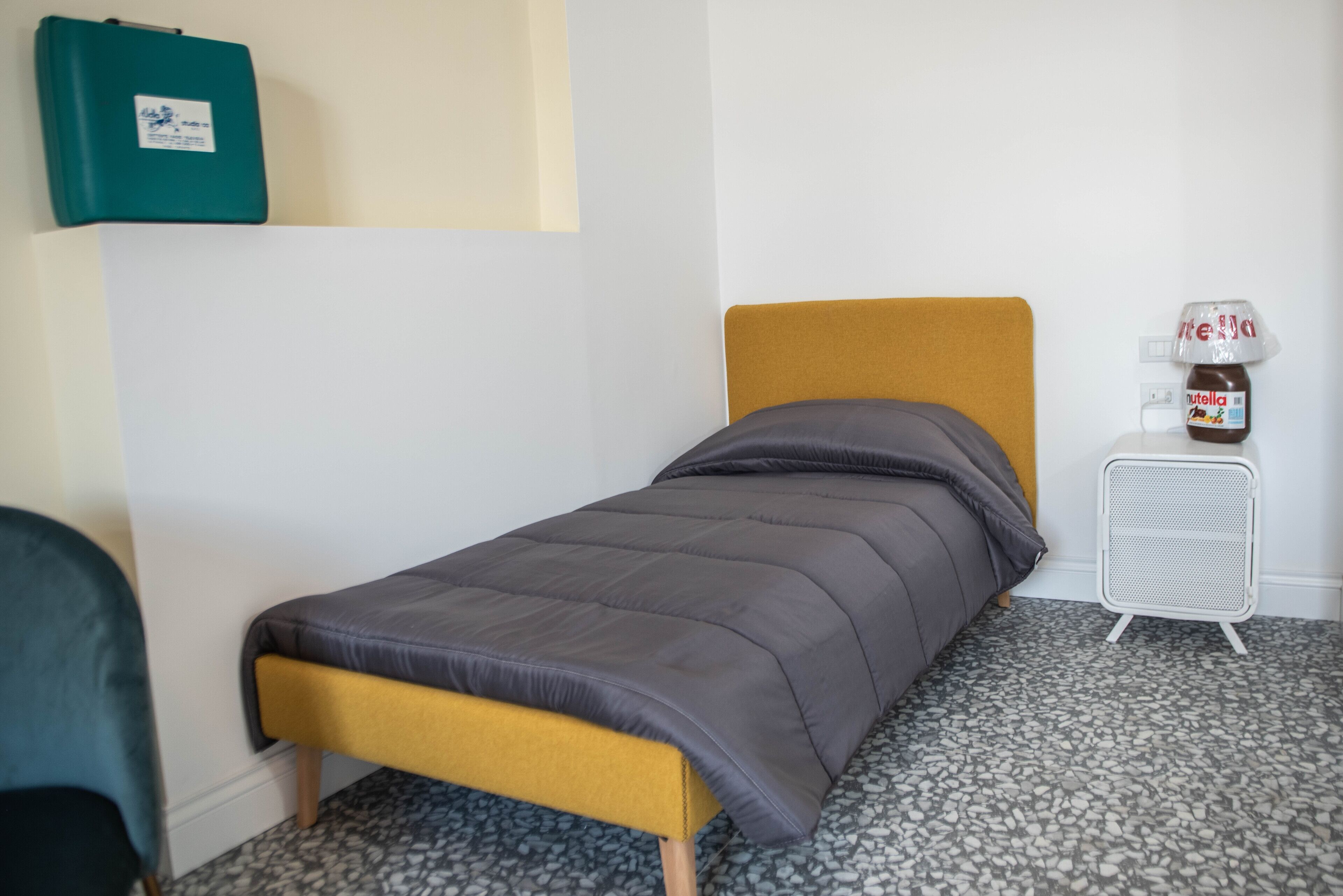 A single bed with a mustard-colored headboard and a gray bedspread in a room with a tiled floor.