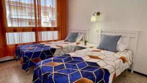 A hotel room with two beds, a window with curtains, a bedside table with a lamp, and a small plant. - OMEGA 4 2-C (Estartit)