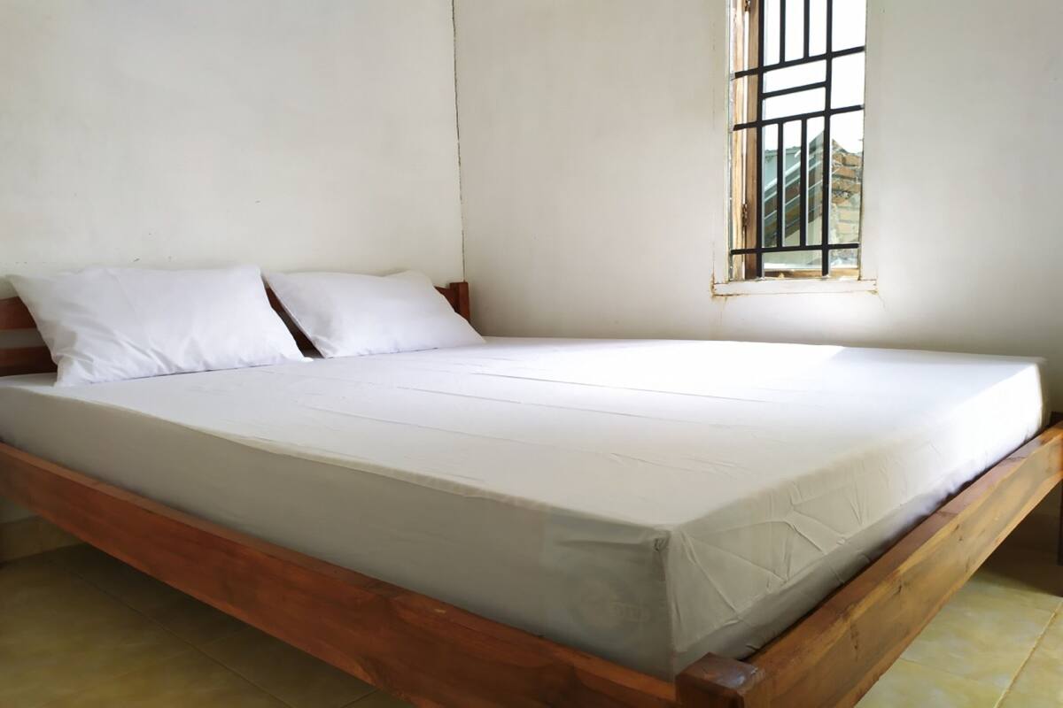 A single-bed room with a wooden frame, white bedding, and a window with bars.