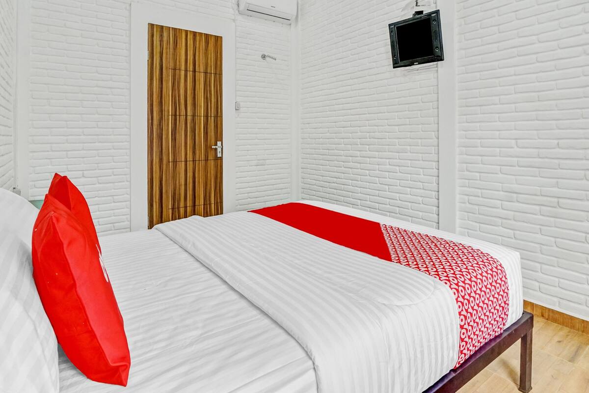Une chambre avec un mur en briques blanches, une porte en bois, un lit avec une literie blanche et rouge, et un petit téléviseur noir fixé au mur.