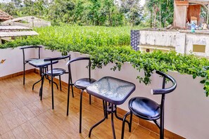 Une terrasse avec quatre chaises en métal noir et une table en verre, entourée d’une végétation luxuriante et d’un mur blanc.