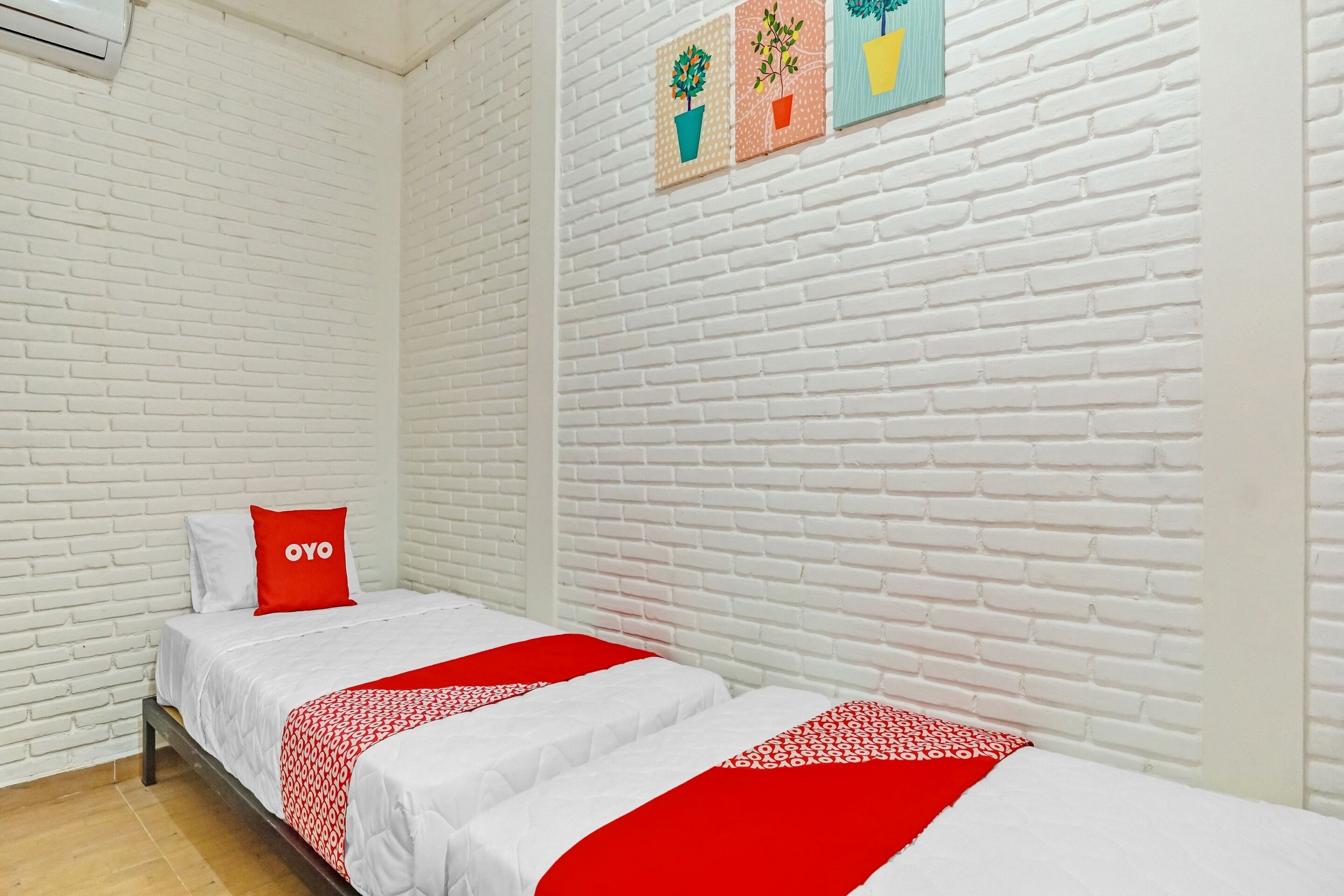 Una habitación con paredes de ladrillo blanco, una cama con ropa de cama blanca y detalles rojos, y tres cuadros colgados en la pared con plantas.