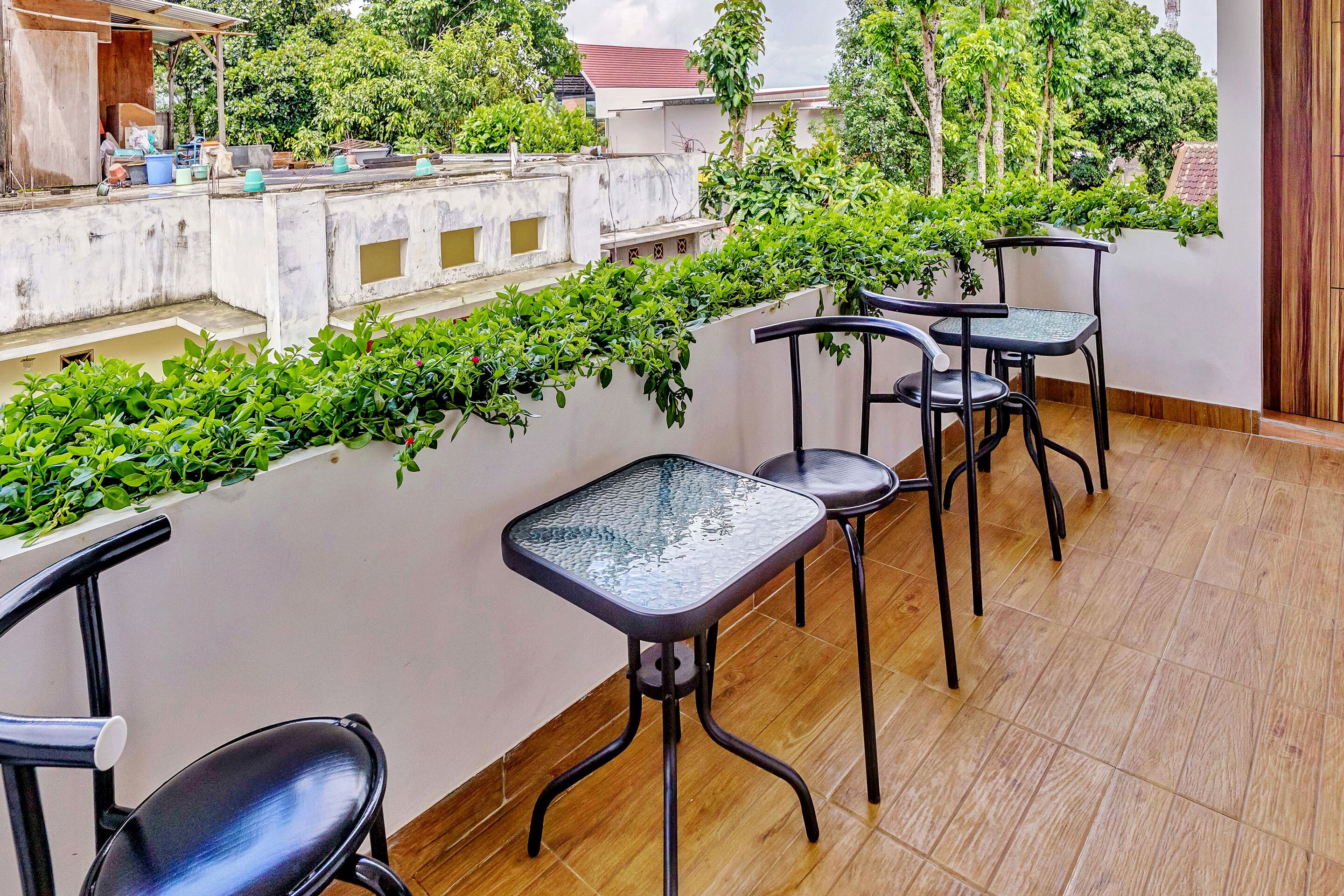 A terrace with a long table and four chairs, surrounded by lush greenery and a view of the cityscape.