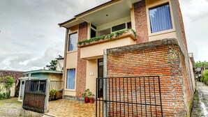 Una casa de dos pisos con balcón, entrada protegida por un portón y un muro de ladrillo.