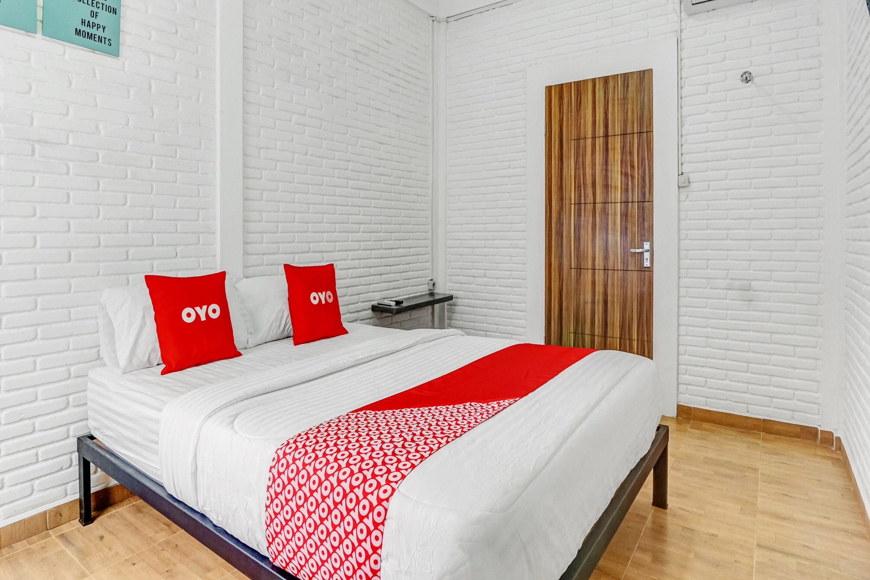 Ein ordentlich eingerichtetes Hotelzimmer mit einer weißen Ziegelwand, einer Holztür und einem Bett mit rotem und weißem Bettzeug.