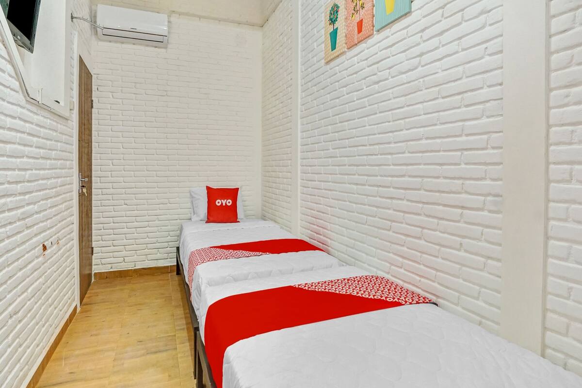 Une petite pièce aux murs de briques blanches, avec deux lits recouverts de draps rouges et blancs, un oreiller rouge orné du mot « OYO » et une petite fenêtre avec un climatiseur.