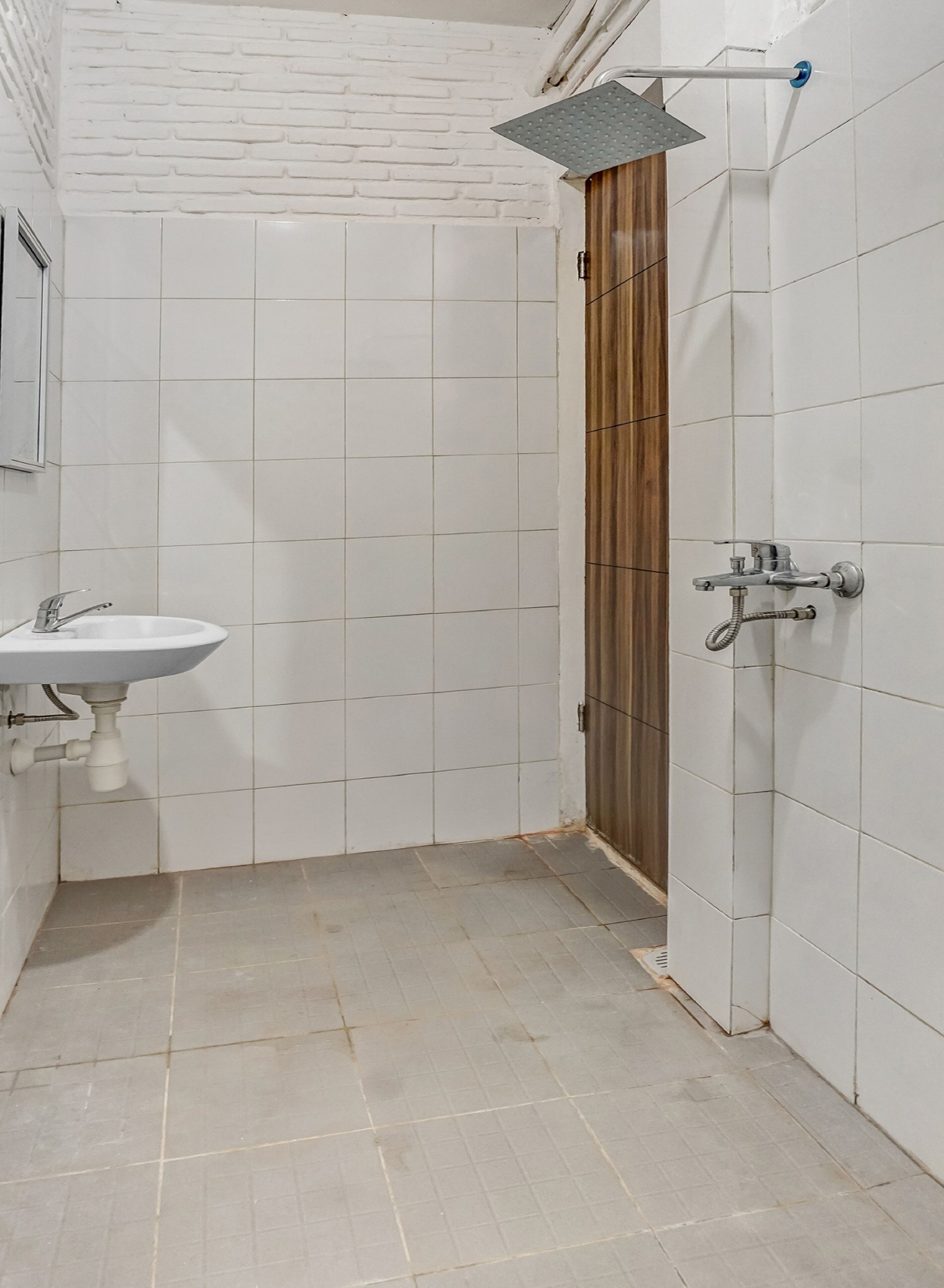 Un baño con paredes de azulejos blancos, una puerta de madera, un lavamanos con grifo y una zona de ducha con ducha.