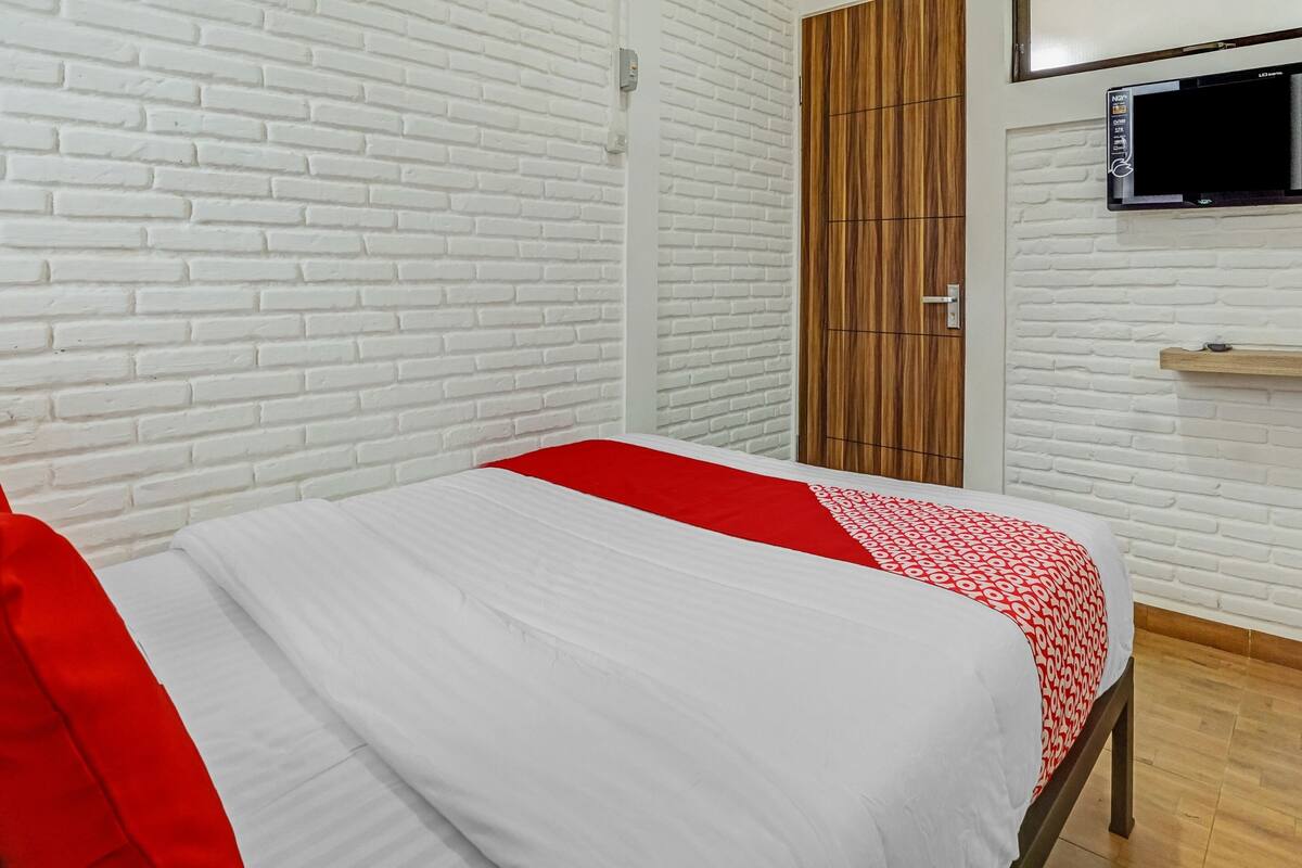 Une pièce avec un mur en briques blanches, un lit avec une literie blanche et rouge, une armoire en bois et un téléviseur fixé au mur.