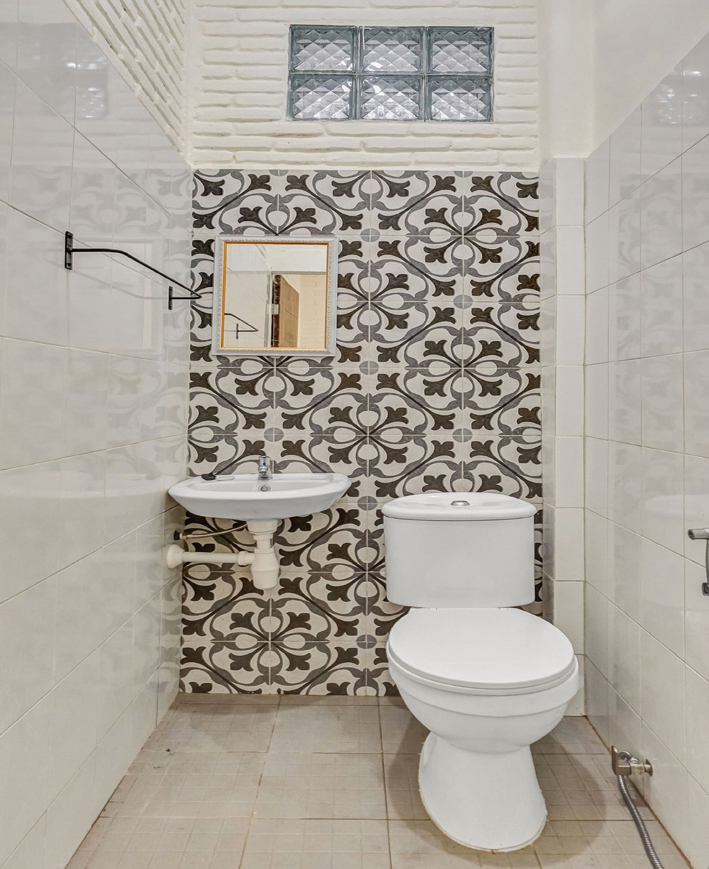 Un baño con pared de azulejos estampados, un lavamanos, un inodoro y una ventana sobre el lavamanos.