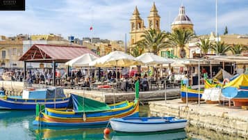 Un puerto deportivo con barcos de colores amarrados, una zona de mercado con sombrillas y edificios históricos al fondo.