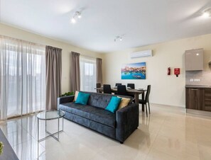 Een moderne woonkamer met een grijze bank, een glazen salontafel en een eethoek met een tafel en stoelen. Er hangt een schilderij aan de muur en een airconditioner bevindt zich boven de eethoek.