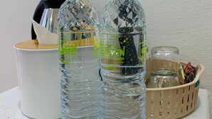 Two clear plastic water bottles with green caps and labels, placed on a white surface next to a white mug and a wicker basket containing various items. - Suanvadee Resort (Koh Samui)