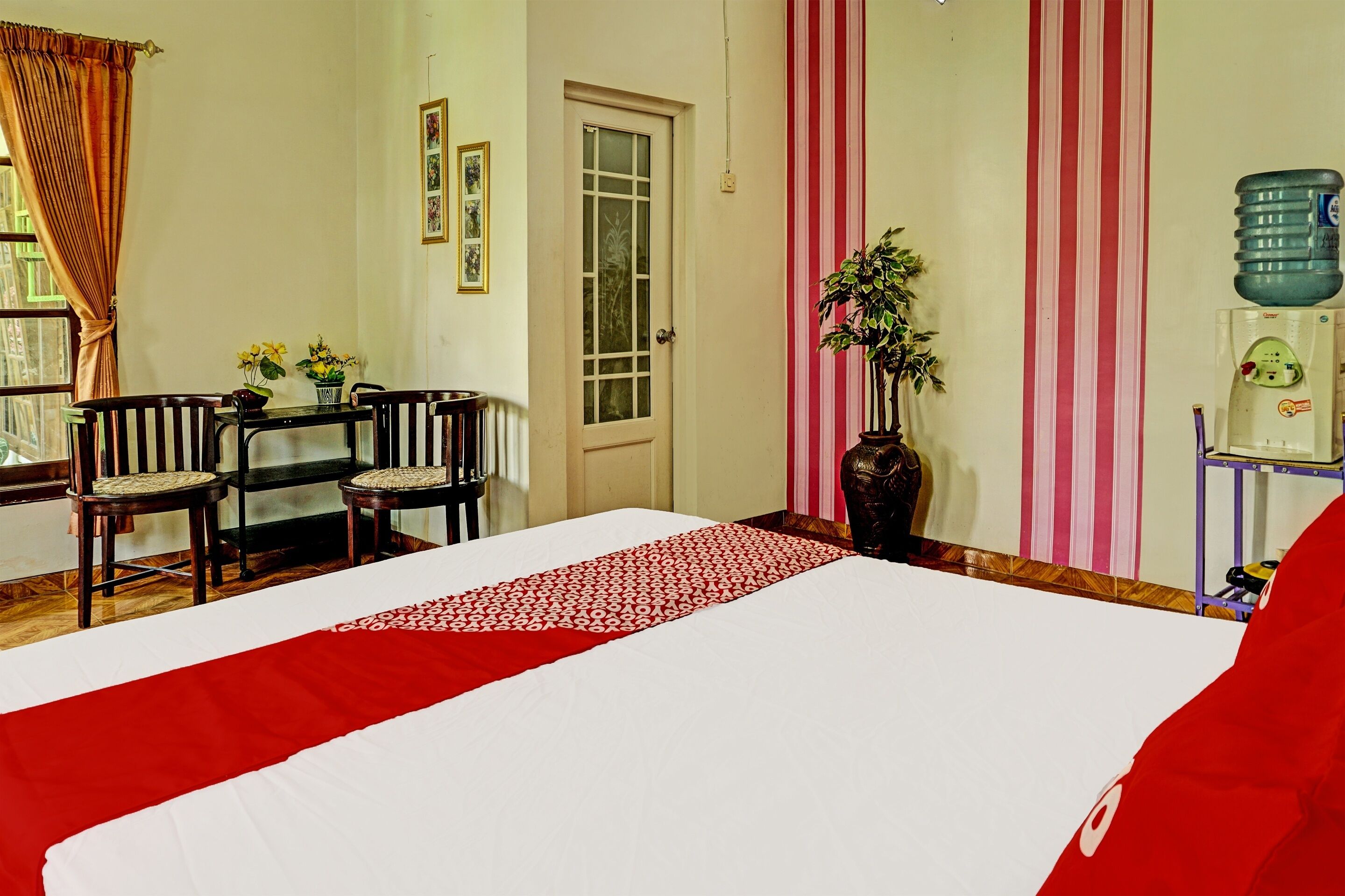 Habitación de hotel con cama, una mesa de comedor con sillas, una planta en maceta y un dispensador de agua.