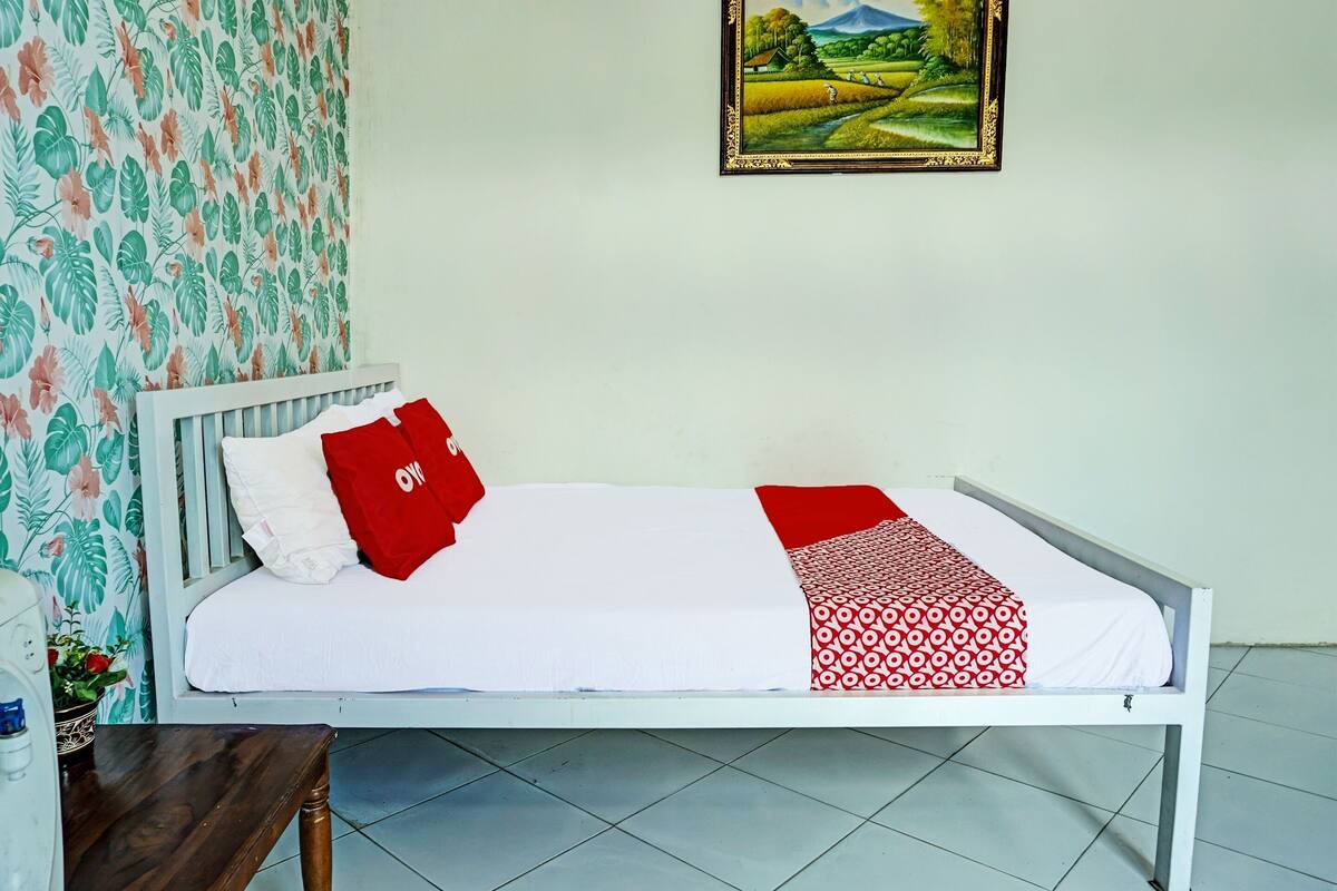 Quarto individual com uma colcha branca, travesseiro vermelho e manta estampada. Há um quadro com uma paisagem na parede.