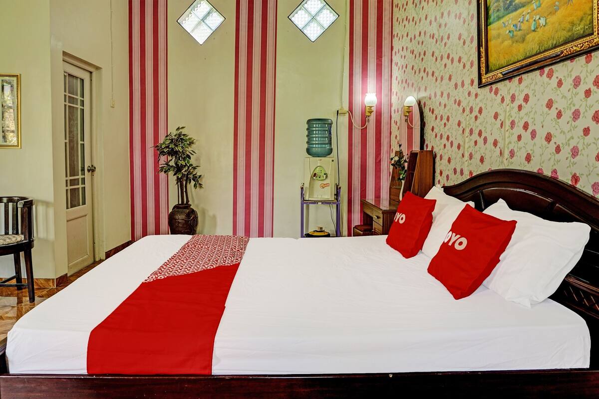 Uma cama bem arrumada com um cobre-leito vermelho e branco, um dispensador de água e um espelho na parede.
