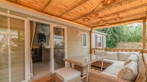 A covered outdoor area with a glass door leading to a kitchen, a wicker sofa, and a glass table. - Mobile Home "Les Pieds Dans L'Eau" with Private Terrace, Private Garden & A/C (Canet-en-Roussillon)