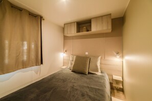 Un dormitorio con cama, mesita de noche, estante sobre la cama y ventana con cortinas.