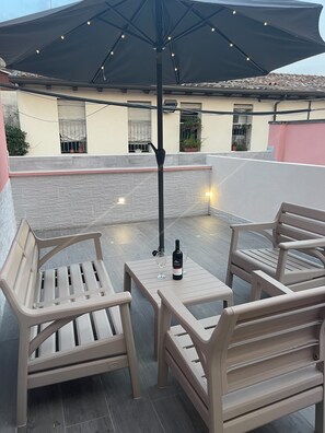 Um pátio externo com mesa, duas cadeiras, uma garrafa de vinho e um guarda-sol grande.