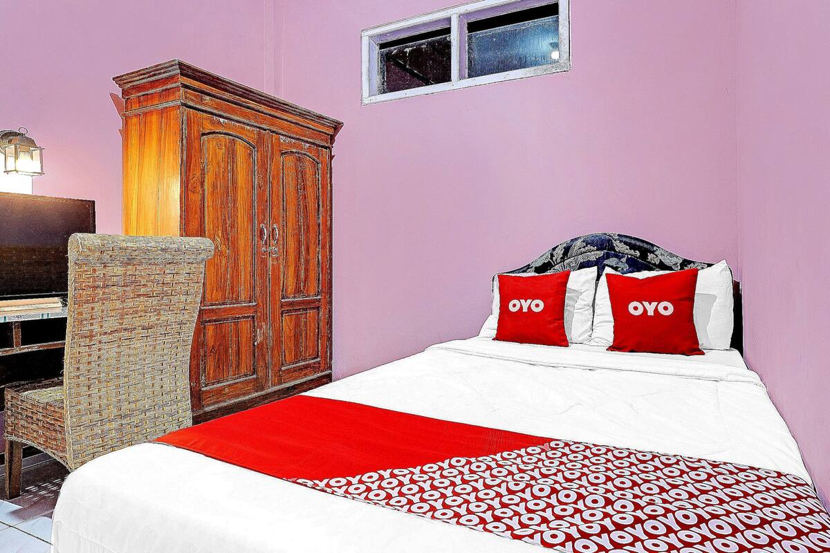Une chambre avec une armoire en bois, une chaise en osier, un lit avec une literie rouge et blanche, et une petite fenêtre.