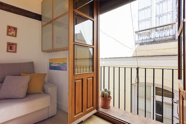 Ein Zimmer mit Holztür, einem Topf-Kaktus, einer Couch mit Kissen und einem Balkon mit Metallgeländer.