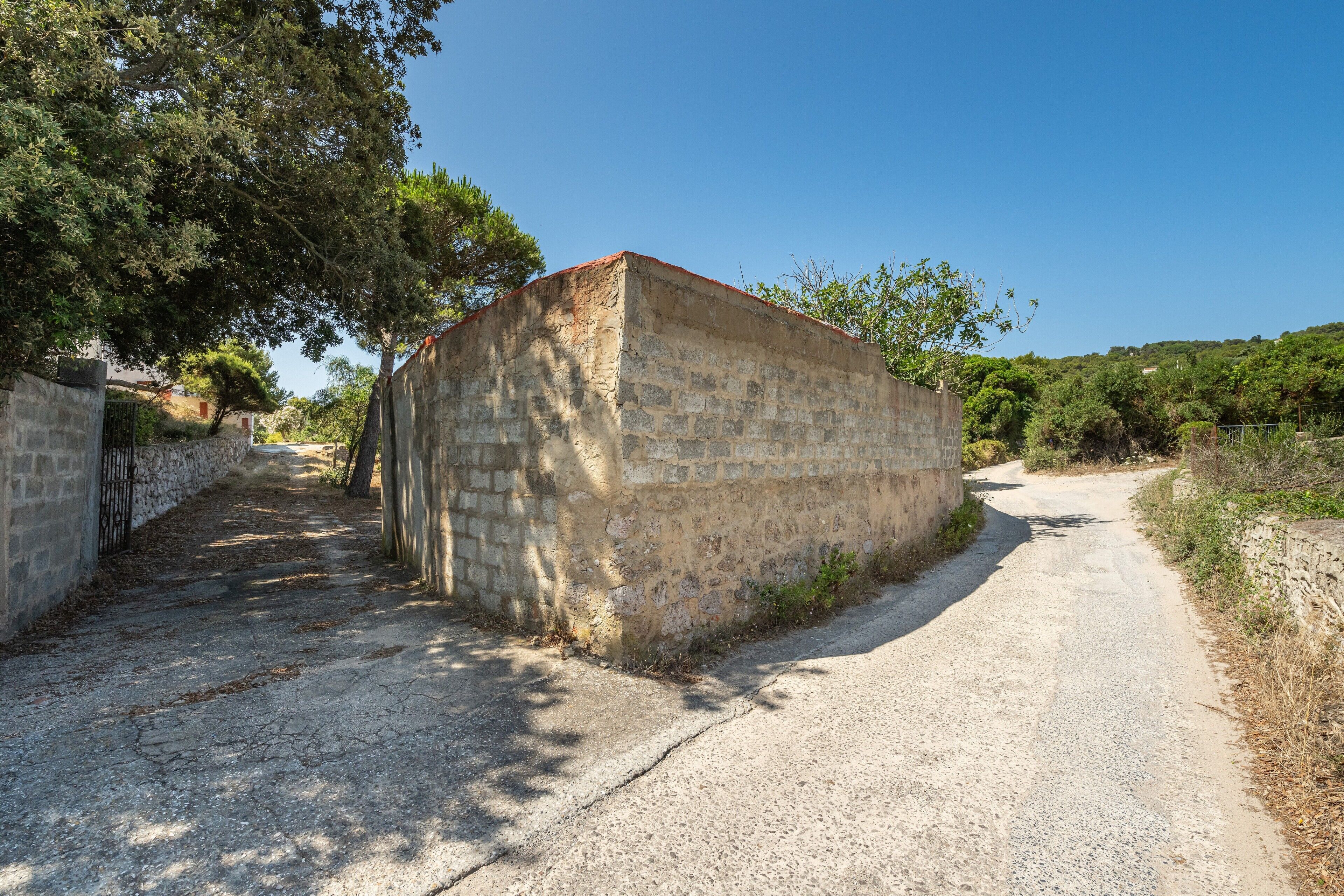 Un camino estrecho y sin asfaltar que conduce a un edificio de piedra con techo plano, rodeado de árboles y vegetación.