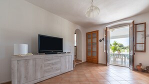 A room with a TV on a wooden cabinet, a ceiling light, and a view through an open door to an outdoor area with a table and chairs. - Holiday Home 'Cala Lunga' with Private Terrace and Private Garden (Carloforte)
