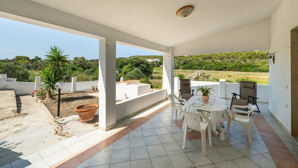 A covered patio with a table and chairs, a potted plant, and a view of a green landscape. - Holiday Home 'Cala Lunga' with Private Terrace and Private Garden (Carloforte)