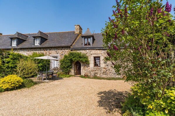 Un bâtiment en pierre avec une allée en gravier, un espace extérieur couvert aménagé en coin salon et un jardin agrémenté de plantes à fleurs.