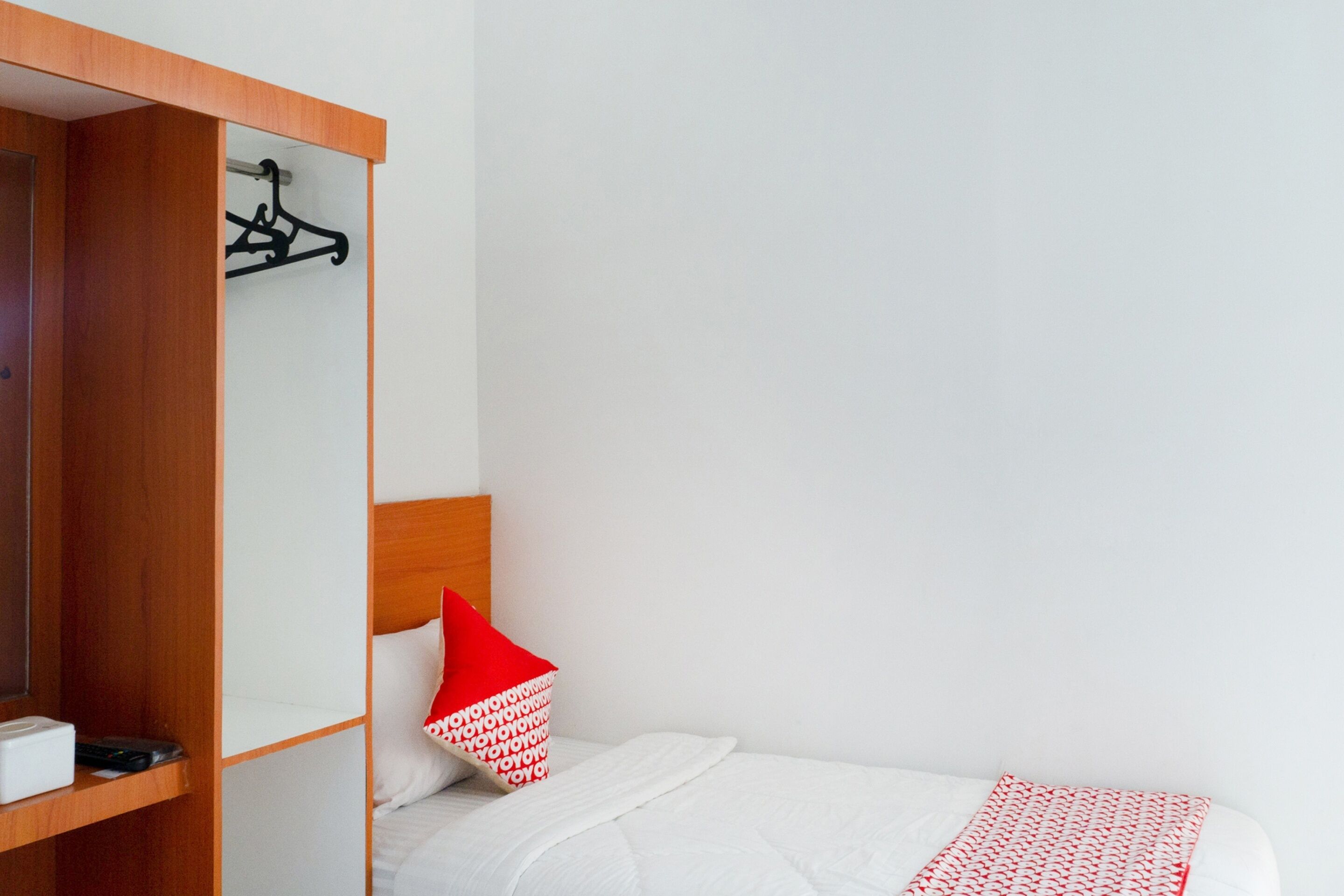 Ein Hotelzimmer mit einem Bett, einem roten Kissen, einer weißen Decke, einem Holzschrank und einem schwarzen Kleiderhaken an der Wand.