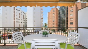 Un balcón con una mesa y sillas blancas, una planta en maceta y vistas de edificios altos.