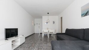 Una sala de estar moderna con un televisor de pantalla plana sobre un mueble blanco, un sofá gris oscuro, una zona de comedor con una mesa y sillas blancas, y piso de baldosas.