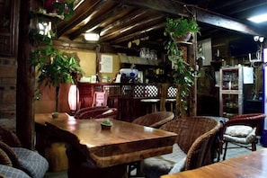 A rustic dining area with a wooden table and wicker chairs, surrounded by hanging plants and a brick wall.