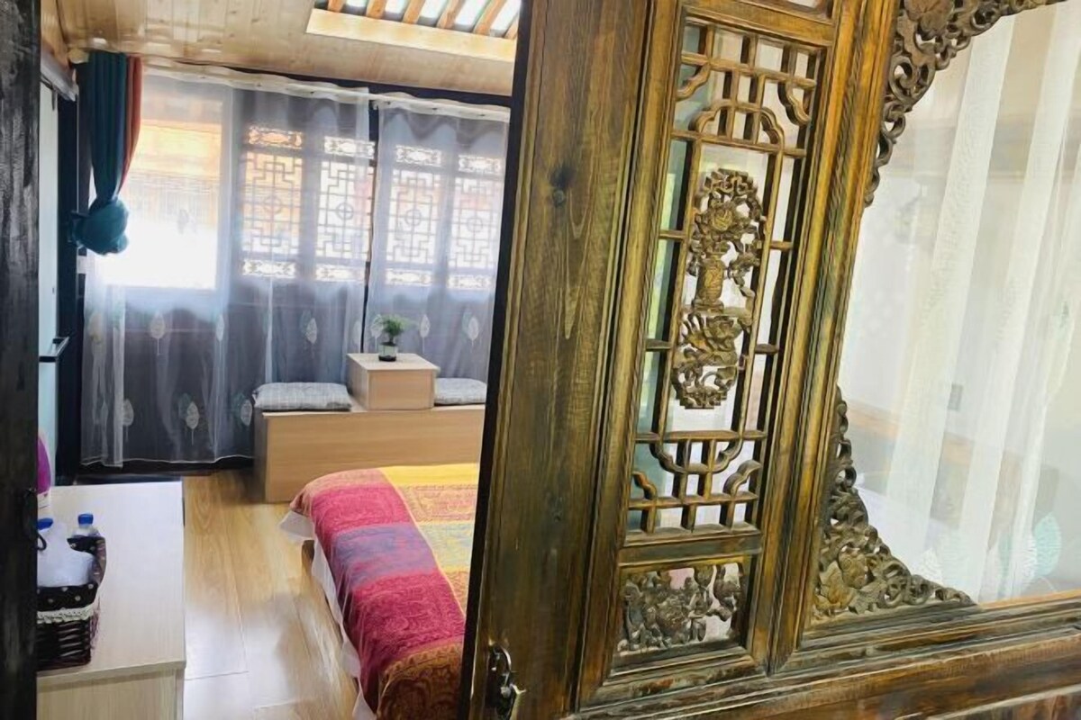 Une chambre traditionnelle avec une porte en bois, un lit recouvert d’une couverture colorée, une petite table avec une plante et une fenêtre ornée de rideaux à motifs.