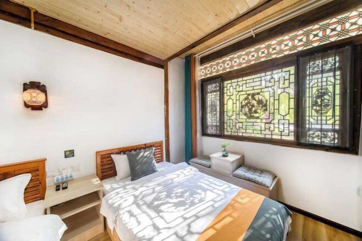 Ein Schlafzimmer mit Holzboden, einem Bett mit gemusterten Betttextilien, einem Nachttisch mit Lampe und einem Fenster mit dekorierter Scheibe.