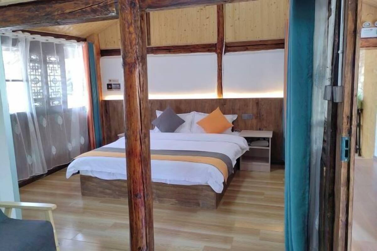 Ein Schlafzimmer mit einem Holzkopfteil, einem Bett mit weißer Bettwäsche und orangefarbenen Kissen, einem Nachttisch und einem Fenster mit durchscheinenden Vorhängen.