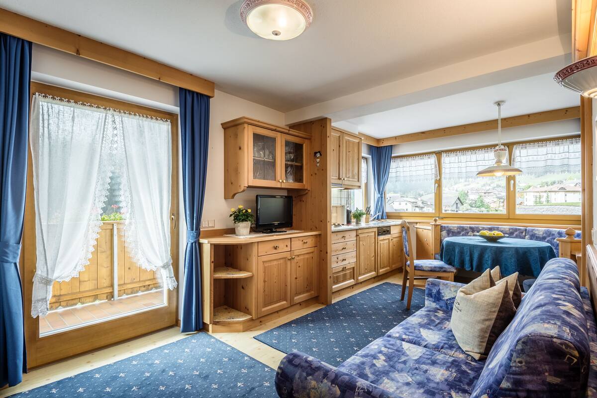 Una habitació acollidora amb un sofà, una televisió, armaris de fusta, una taula de menjador i una finestra amb cortines.