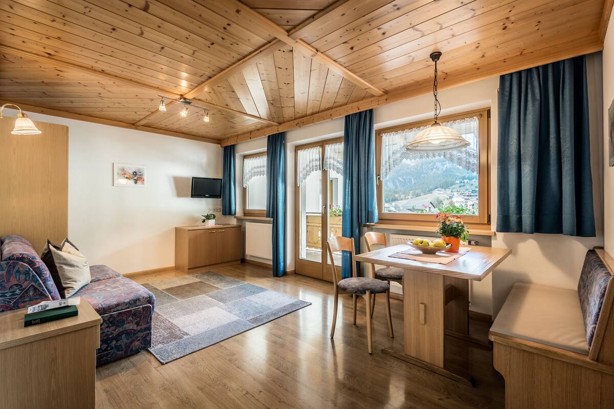 Una habitació acollidora amb un sostre de fusta, una taula de menjador amb cadires, un sofà i un televisor penjat a la paret.