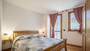 A bedroom with a wooden bed, a nightstand, a lamp, and two windows with curtains. - Holiday Apartment "Agritur Piazera Con Terrazzo" with Mountain View (Ronzo Chienis)