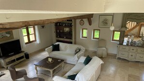 A spacious living room with a white sofa, a brown armchair, a coffee table, and a television. There are bookshelves, a chandelier, and a staircase visible in the background. - Maison Solognote au Coeur des Châteaux de la Loire (Tour en Sologne)