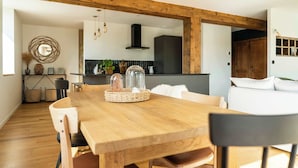 A wooden dining table with chairs, a kitchen in the background, and a white sofa with pillows. - Gîte d'exception avec vue panoramique (Prémanon)