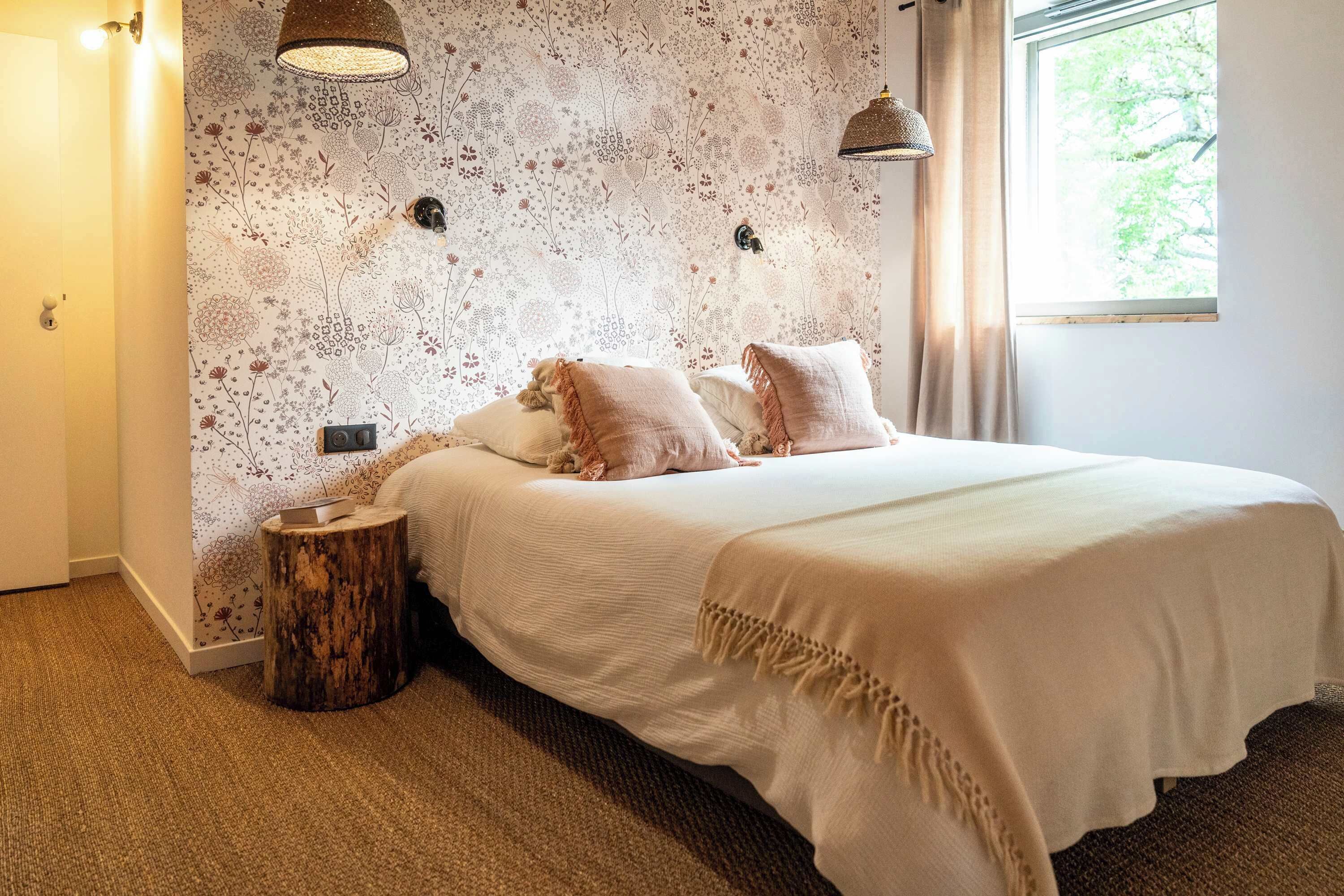 Un dormitorio con papel tapiz floral, un taburete de madera, una cama con ropa de cama blanca y dos lámparas colgantes.