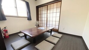 A room with a wooden table, four cushioned seats, a fan, a fire extinguisher, and a shoji screen door. - chidoriasahinoie (Osaka)