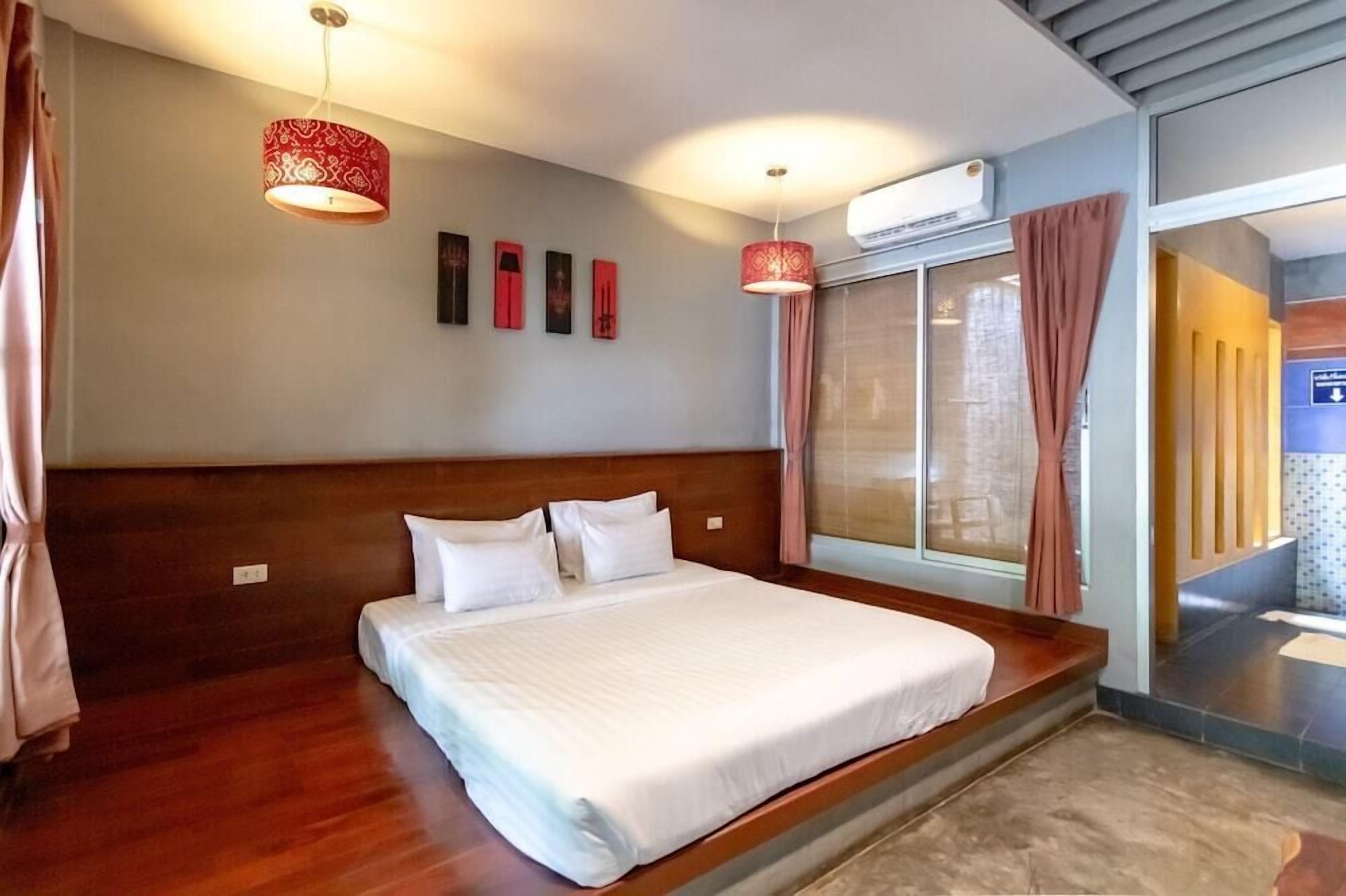 A hotel room with a large bed, a wooden headboard, a window with curtains, a wall-mounted air conditioner, and a hallway with a sign.