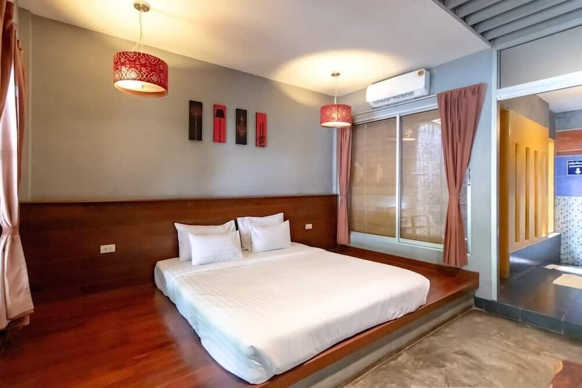 A hotel room with a large bed, a wooden headboard, a window with curtains, a wall-mounted air conditioner, and a hallway with a sign.